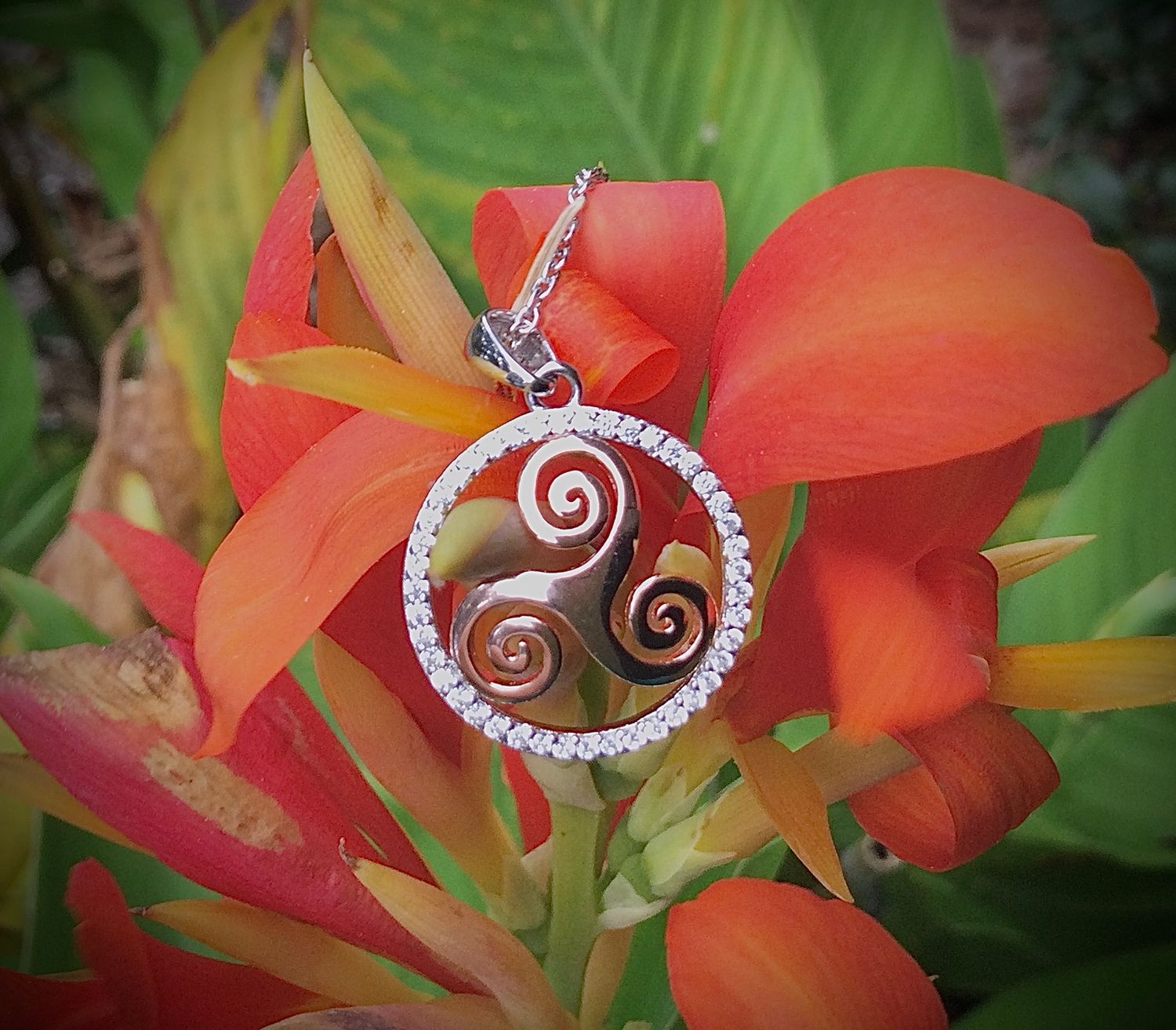 Sterling silver triskele pendant with rose gold plating, surrounded by sparkling cubic zirconia stones, shown on flower. 